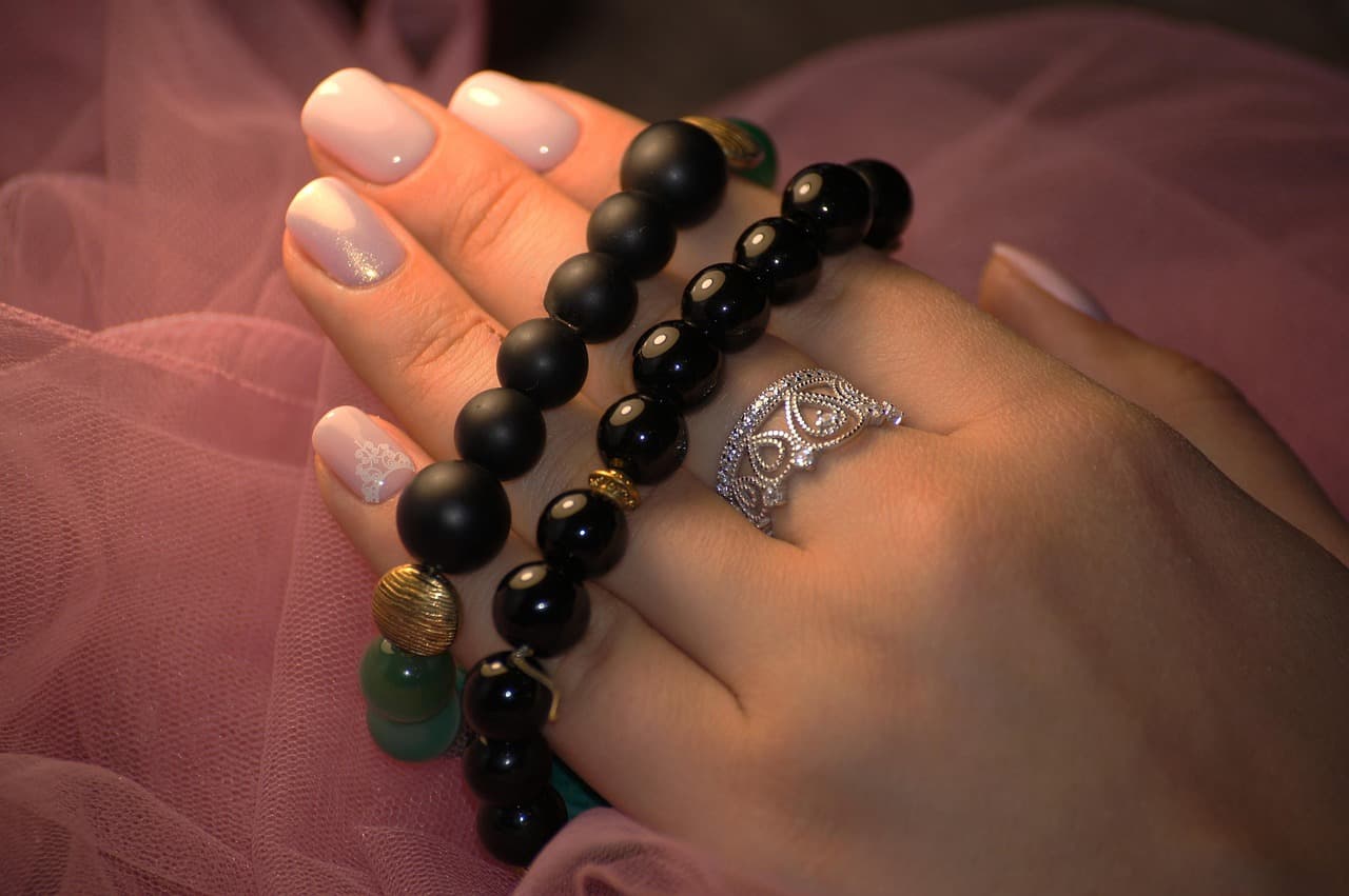 A hand with milky pink nails, one with white lace detail, wearing black bead bracelets and a silver ring, on pink tulle
