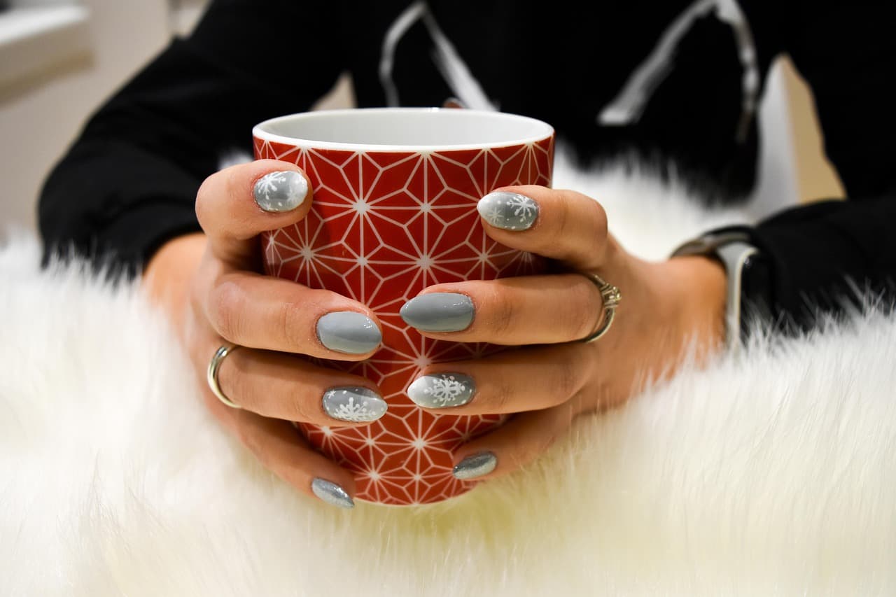 Hands with grey gel nails and silver snowflake designs holding a red patterned mug against white faux fur